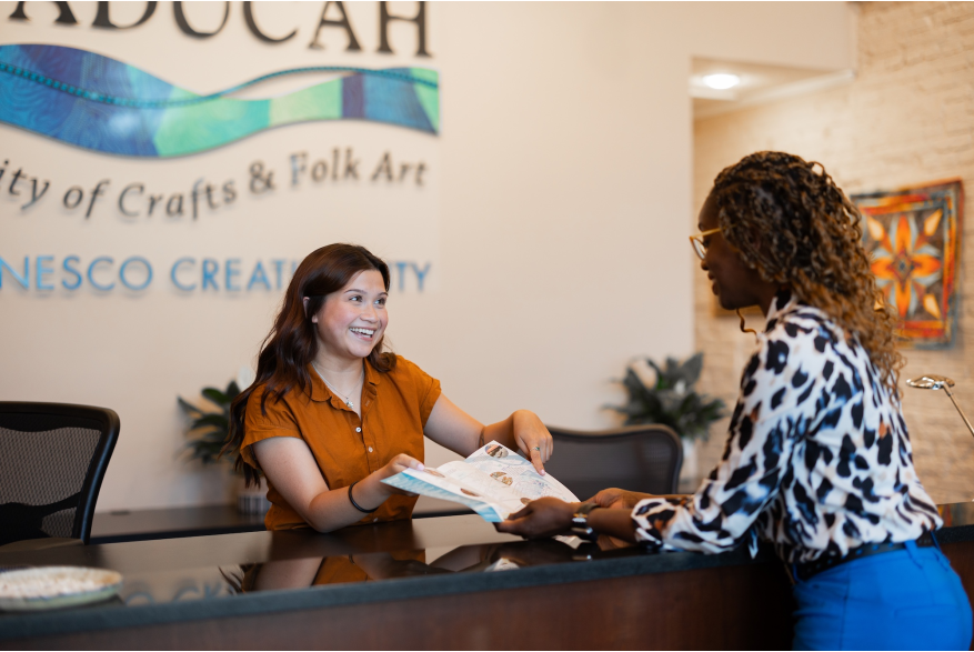 A woman stands behind the counter at the Paducah Convention & Visitors Bureau and helps another woman who is visiting Paducah. They are holding a map and pointing at a location.