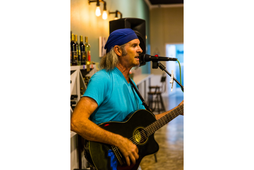 A man in a light blue shirt and dark blue hat strums a guitar and sings into a microphone.