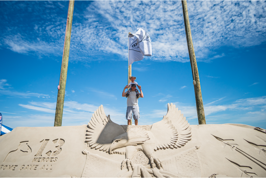 A man carries his toddler daughter on his shoulders and they pose atop a large sand mountain carved with an eagle