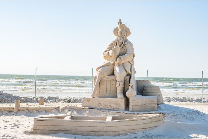 Sand sculpture of a pirate drinking out of a cup and sitting on barrels. In front of him is a rowboat made of sand