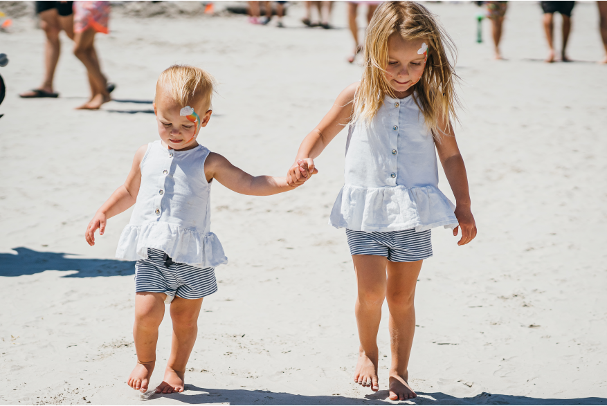 Two toddlers hold hands on the beach in matching outfits. Both of their faces are painted.
