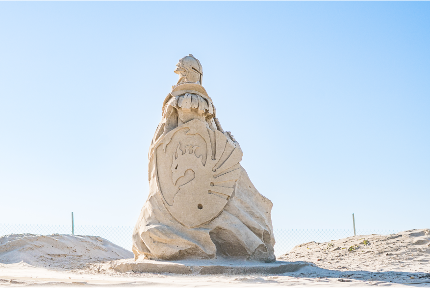 Sand sculpture on the beach of a guardian soldier facing the left. The soldier carries a shield emblazoned with a dragon.