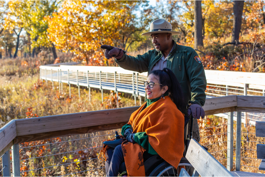 A brunette woman using a wheelchair looks at the fall colors in the distance. A park ranger points to something out of the picture.