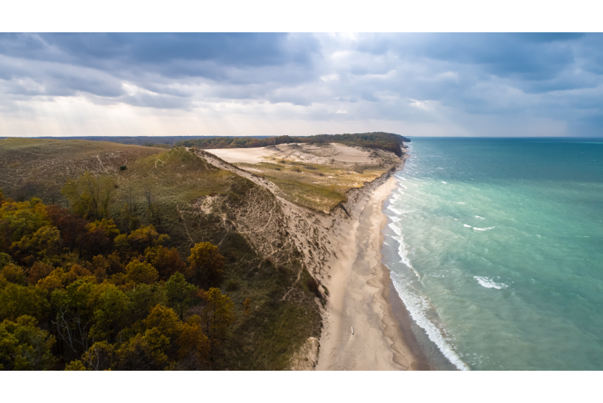 Aerial view of a lakeshore. The water on the right is a variety of light and dark blues. The dunes end in green vegetation and woods.