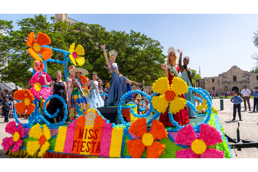 Women dressed in gowns and crowns on a colorful Fiesta float in front of the Alamo.