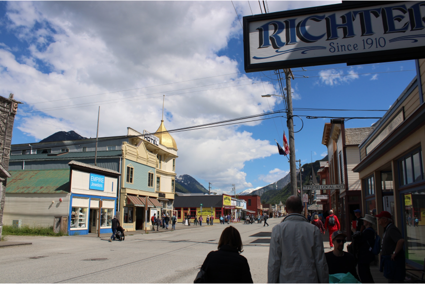 Scattered clouds, blue skies and a gold rush themed western style street scape . Old building with false fronts and a gold domed building. People walk on boardwalks and in the grey asphalt street