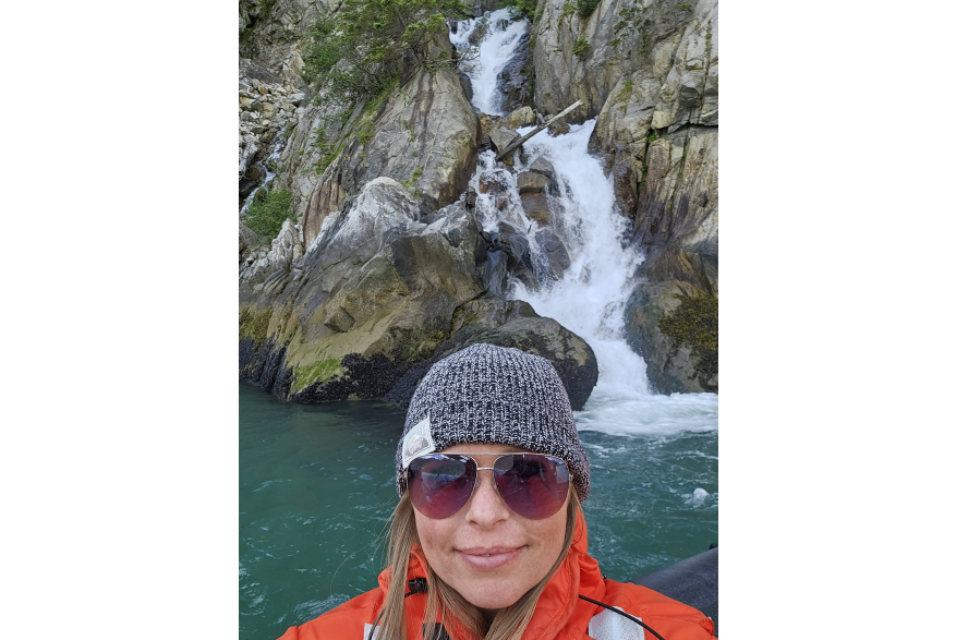 frothy white waterfall cascades down a rocky mountain into the green sea. Woman smile for selfie in front wearing a bright orange Mustang life suit, pink tinted aviator glasses and a black and white fitted beanie over strands of blonde hair