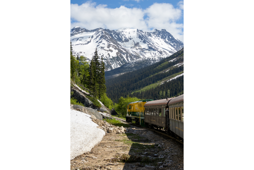 green and yellow locomotive pulls brown train cars through the green tree covered mountainous terrain. Dirty melting snow sits to the left of the tracks. Dark blue snow covered mountains, and partly cloudy blue skies lie beyond.