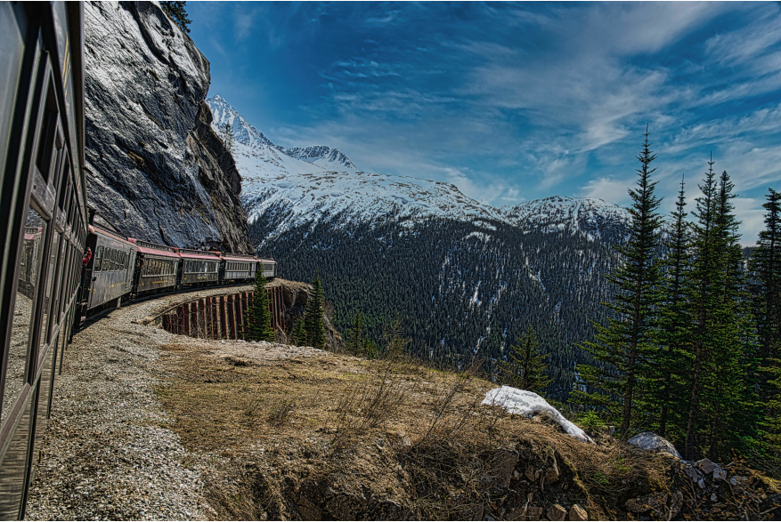 Brown and red roofed train passenger cars Coming around the bend under partly cloudy blue skies blue and white snow capped mountains and ever green trees in the distance