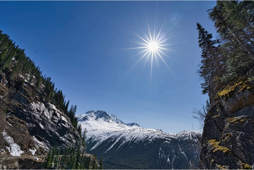 The sun bursting in rays in the clear blue sky with jagged blue and white snow capped mountains in the distance with evergreen trees framing the side views