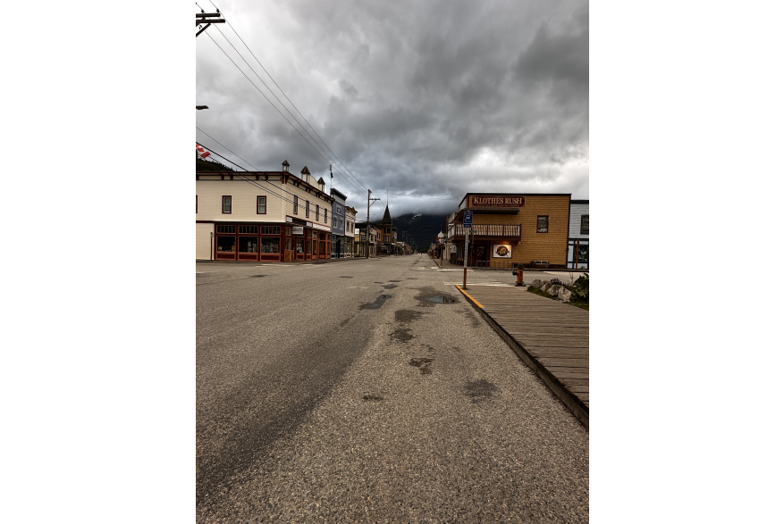 Empty asphalt streets and wooden boardwalk with false front, 1800's Gold Rush style buildings lining each side under cloudy skies and powerlines.