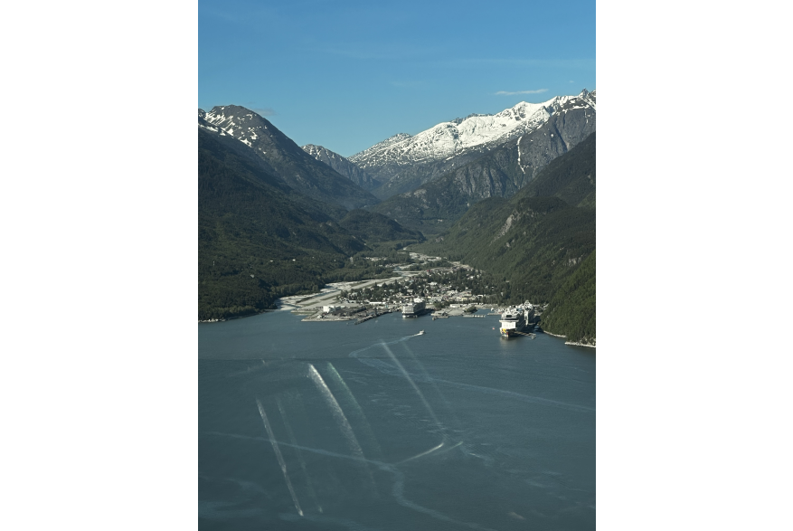 Layers of steep towering mountains from snow capped to evergreen covered mountains dive into a narrow valley with a river on the left and three cruise ships positioned  at fingerlike dock infrastructure