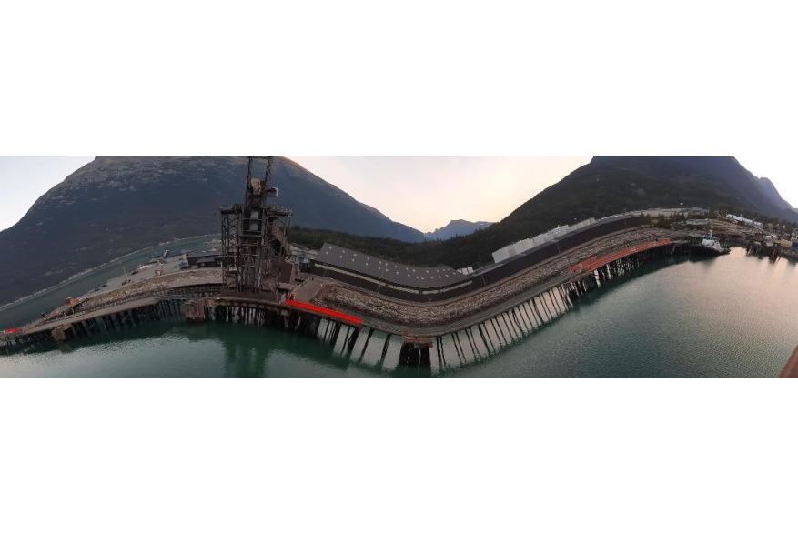 a fish bowl view of the wooden dock and Ore Ship loader and facilities creates an illusion that the structures are bent down towards the still green ocean water below with two dark blue mountain peaks towering into the white evening sky behind it