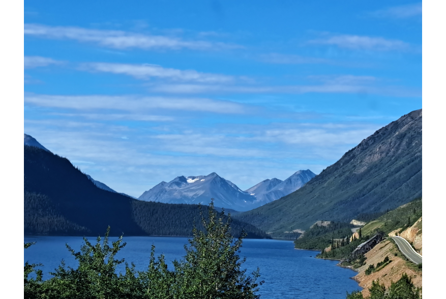 Bright blue skies with scattered clouds over a vast dark blue lake with mountains rising up on either side and off in the distance. Green leafy trees in the foreground. Off to the right and highway winds along the mountain side past an old mine building at the edge of the lake and on into the distance