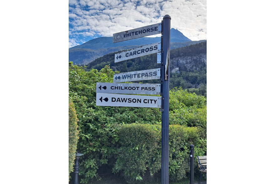 a sign post with white signs jetting out in every directions reads "Dawson City" Chilkoot Pass "White Pass" Carcross "Whitehorse. groomed bushes and a park bench lie just beyond. In the distance mountains rise high into the bright blue sky scattered with white clouds.