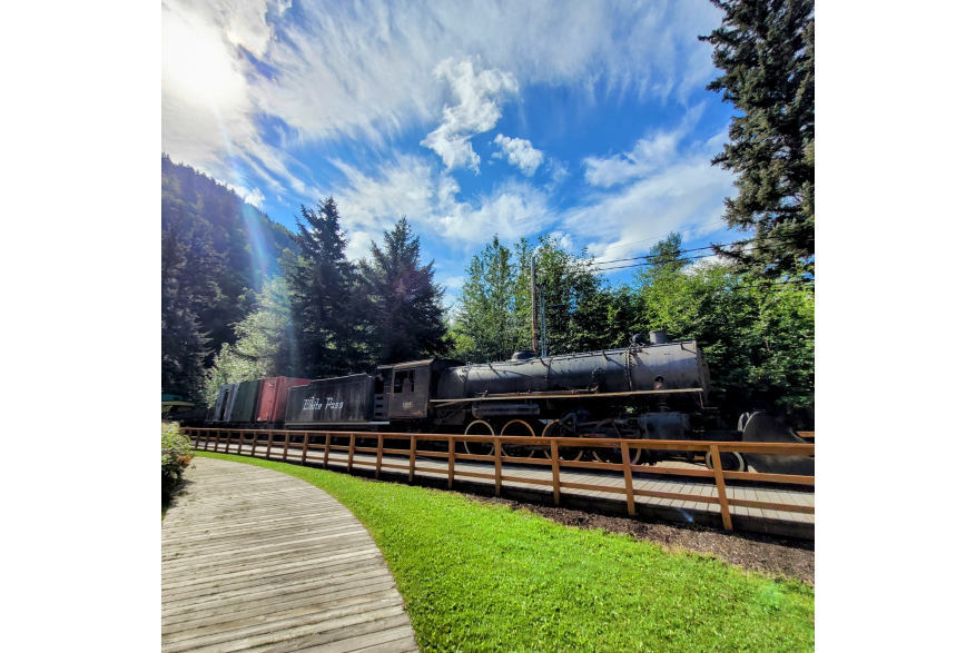 Sun streams out from scattered clouds in a bright blue sky. A black steam locomotive sits on display behind a neat wooden fence and boardwalk with a sea of green ever greens and leafy trees behind it.