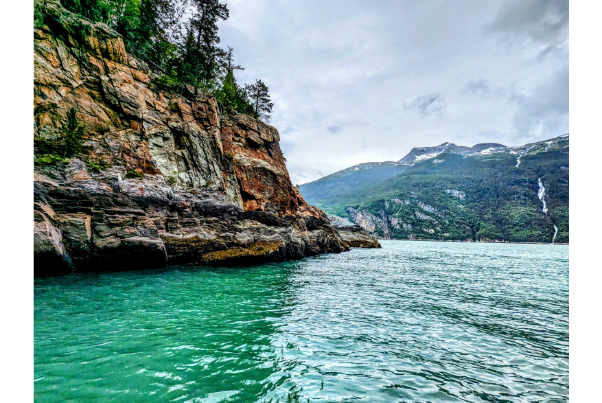 photo taken from the blue green seascape with grey gold cliffs to the left and more ocean up ahead. A white waterfall cascades through the evergreen covered mountains with light grey cloud covered skies overhead