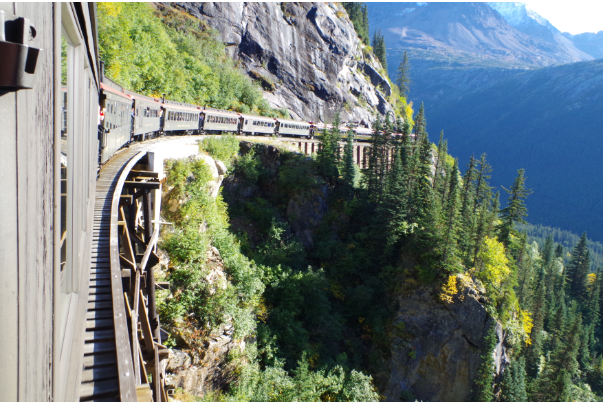 Brown train cars with red rooftops travel over narrow gauge railway over a wooden bridge and around a cliff hanging turn through mountainous green forest terrain White Pass & Yukon Scenic Train Ride