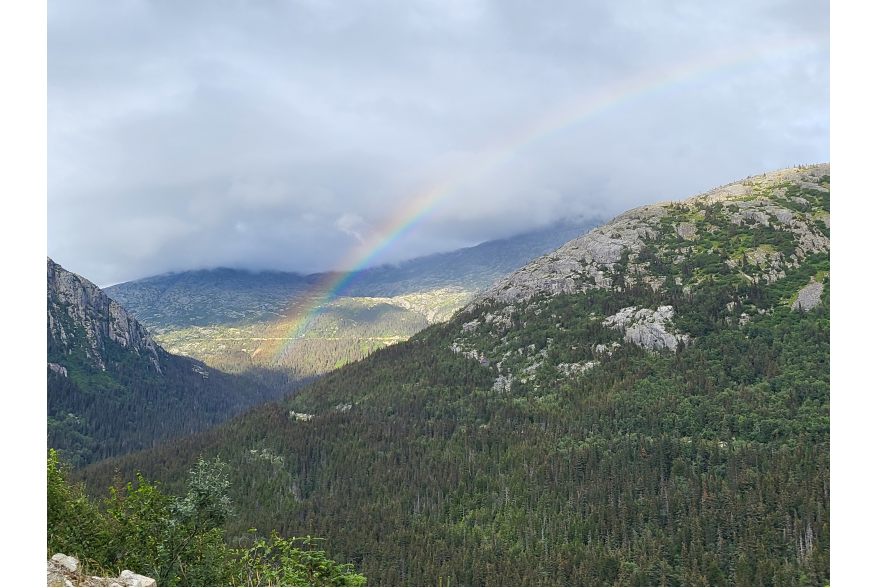 a rainbow under cloudy skies arches over rocky evergreen covered mountains and dives into a valley