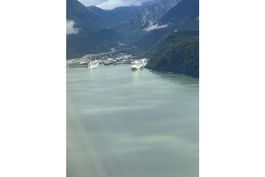 an aerial shot of the Light green ocean water is patterned in shadows from the spotty cloud cover overhead. The waters but up against a narrow valley full of small buildings. Three giant white cruise ships are parked on either side of the town. Massive evergreen covered mountains rise up from the valley on either side in into the distance ahead, the mountains grow taller and snow covered.