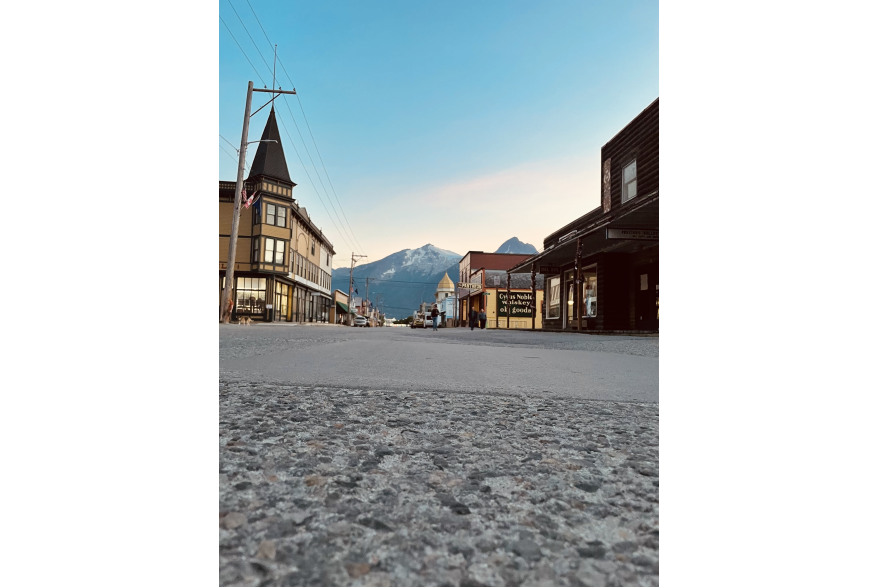 aggregate asphalt in the foregroud gives rise to gold rush era false front buildings on either side of a virtually empty Broadway street with massive snow topped mountains in the background a blue skies at dusk
