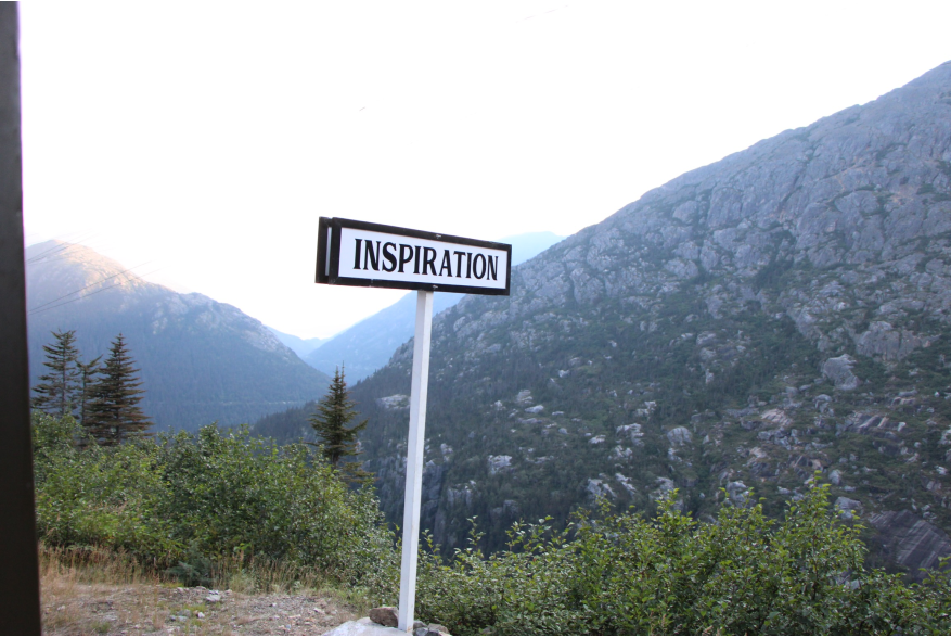 I tall white sign post with a long white sign with black border and block lettering reads "Inspiration". Evergreen and bright green deciduous trees lay behind it, dipping into a canyon. In the background at varying distance are three mountains rising out of the valley with bright white skies overhead.