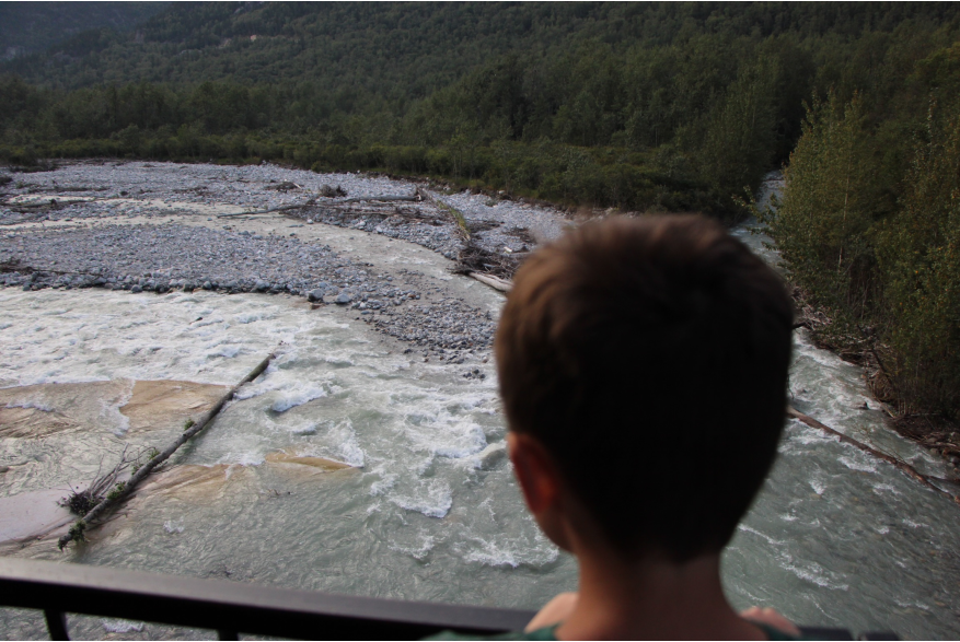 a little boy with short brown hair gazes out the into the vast Alaskan wilderness from the back deck of a train car laying eyes on a white water frothing river raging over river rock and through evergreen trees. Several logs line the path of the river bed from trees that have succum to mother nature