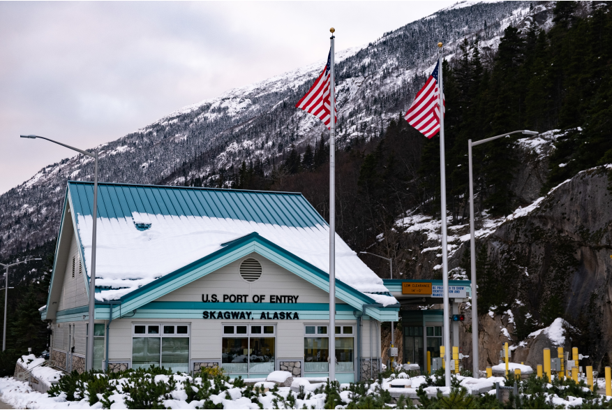 US Port of Entry grey building with blue roof half covered in snow with two American flags on poles in frontSkagway border crossing station with Porcupine Hill behind