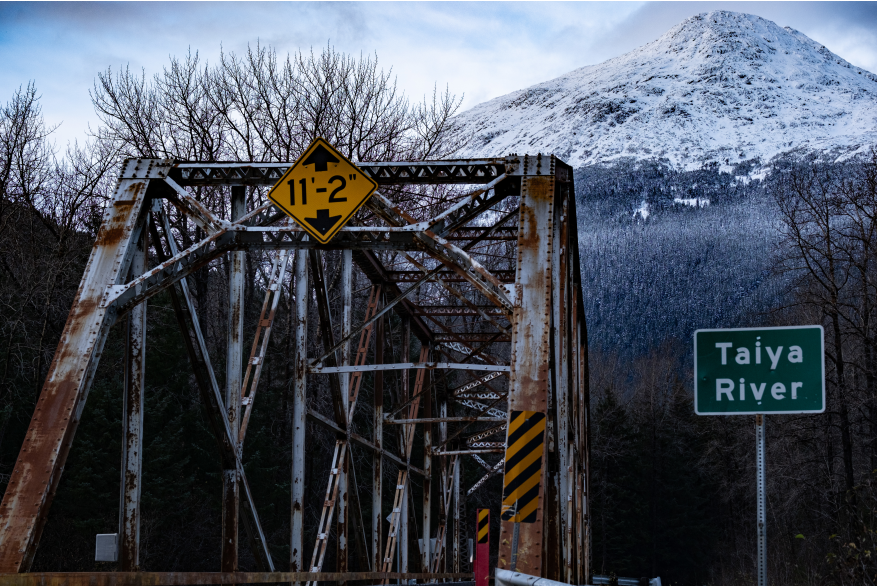 Rusty steel bridge with yellow traffic sign warning max height of 11'2' to cross Tayia River with snowy mountain peaks and leave barren trees in the background at the Chilkoot trailhead