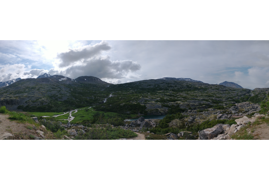 Panoramic view of white waterfalls emerging from massive rolling rocky mountain hills into a lush green valley as the sun shine breaks out from behind fluggy white clouds