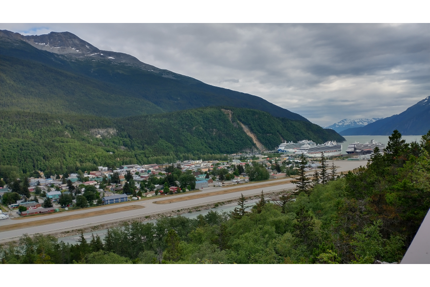 Vantage point of Skagway from the Lookout on Dyea Road show a long narrow airstrip and townsite nestled perfectly in the valley floor with giant evergreen covered mountains steeply jetting up behind it. Three Cruise Ships sit iddle on calm dark seas.