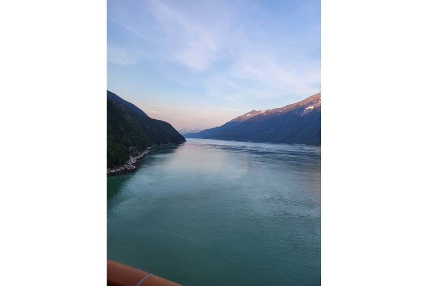 The wooden rail of a cruise ship deck is barely visible in the left corner of the photo that gives way to lightly roughed up dark blue green waters between two steep evergreen covered jagged mountain ranges in North America's deepest fjord under pink and blue sunrise skies