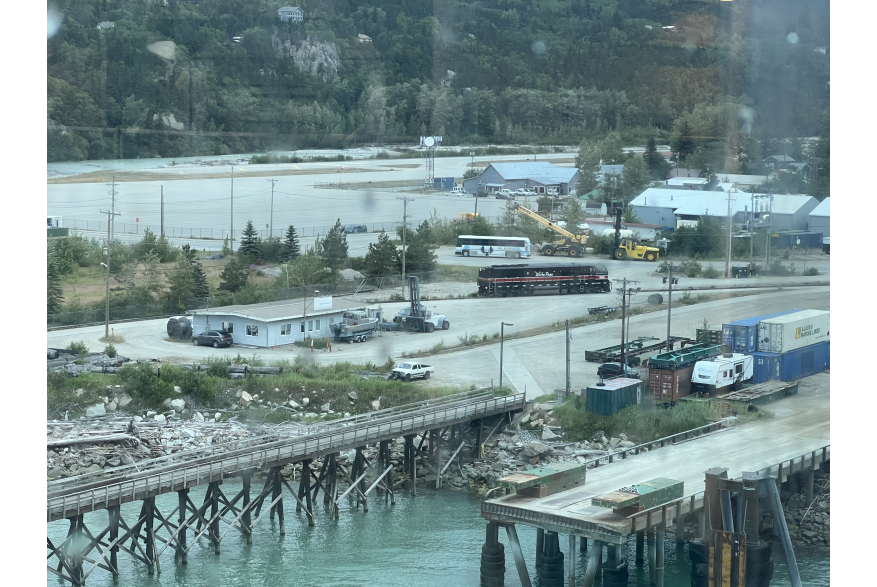 Photo through water spotted glass of freight dock and Ore Dock with Black and Red locomotive in the industrial uplands of Skagway. shipping containers, motorcoach, cars and buildings. blue green oven in the forfront and evergreen covered mountains in the background