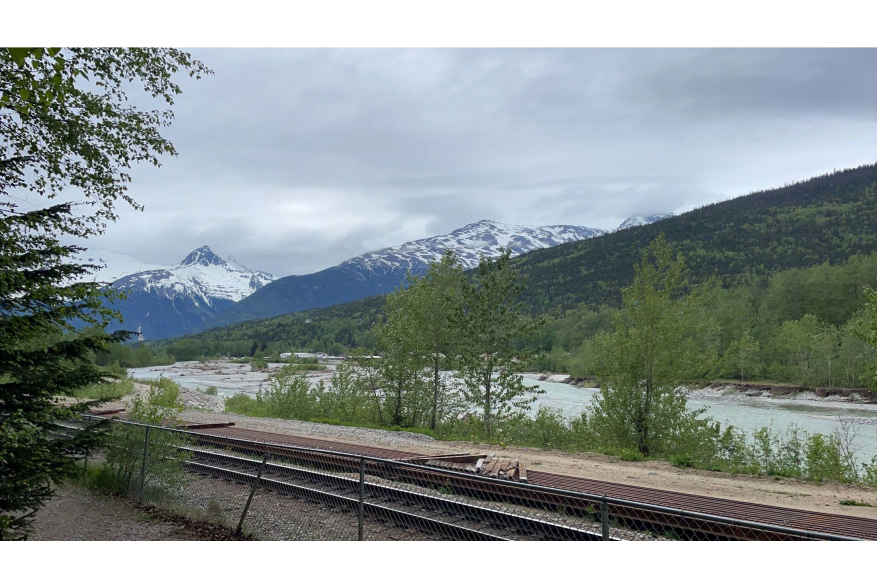 chain link fence in front of railroad tracks with Skagway River flowing in the background and green trees and blue snow capped jagged mountain peaks in the distance under grey cloudy skies