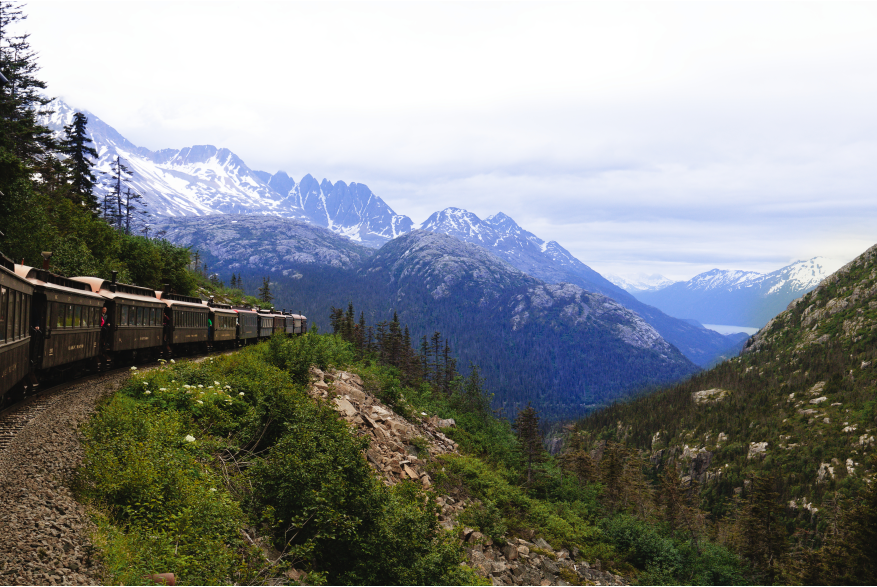 A line of vintage train cars curves along the mountainside, cutting through forested and rocky terrain. Snow-capped peaks rise over the White Pass and Yukon Route in Skagway.