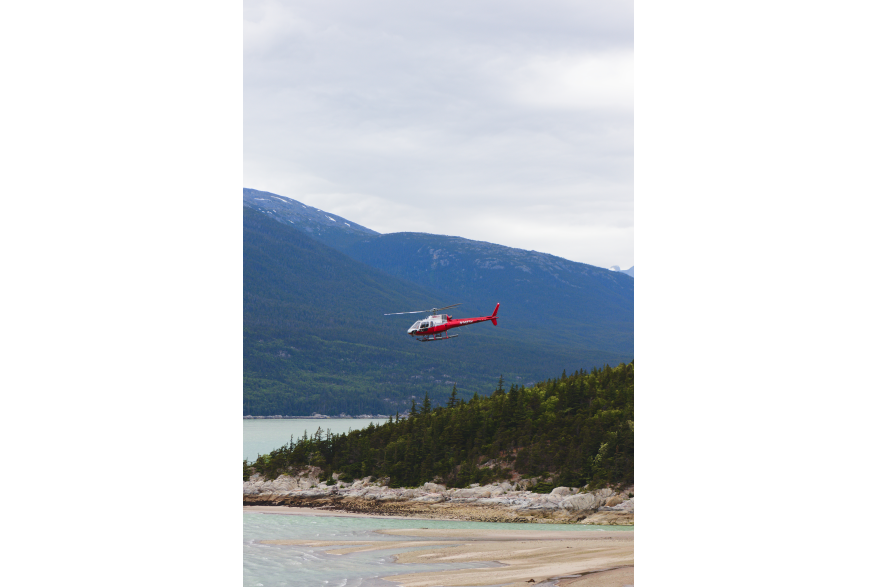 A red helicopter flies over Skagway’s rocky shoreline and forest, with mountains rising in the background under a gray sky. The light is soft, bringing out the textures in the trees and the turquoise water.