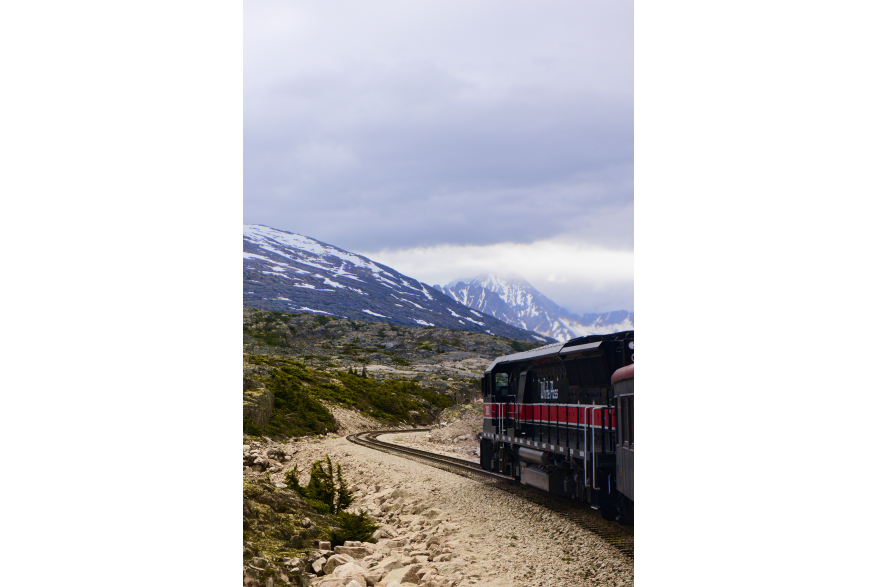 A train on the White Pass and Yukon Route curves along the track through rugged terrain in Skagway, Alaska. Snow-covered mountains rise in the background under cloudy skies.