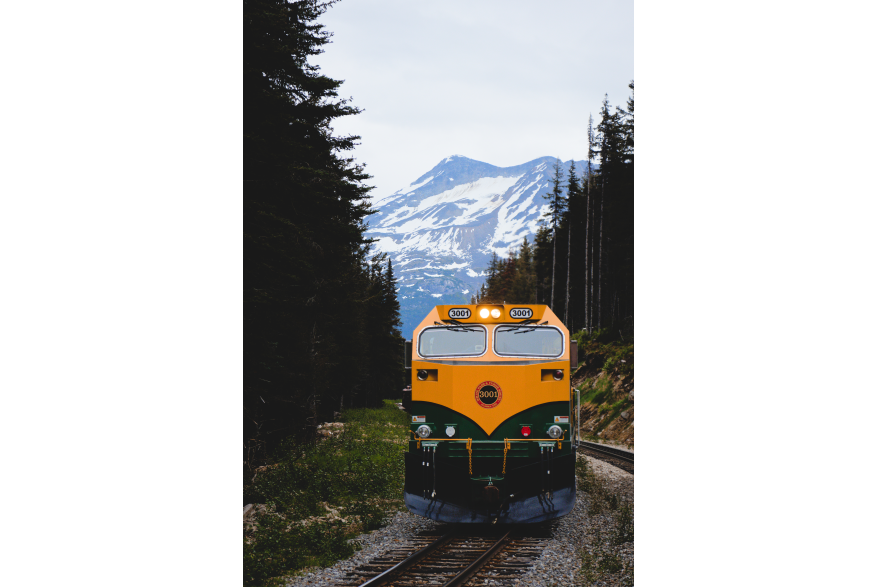 A yellow and green locomotive from the White Pass and Yukon Route drives between pines with snow-covered mountains behind. The train lights shine under an overcast sky, contrasting with the Skagway landscape.