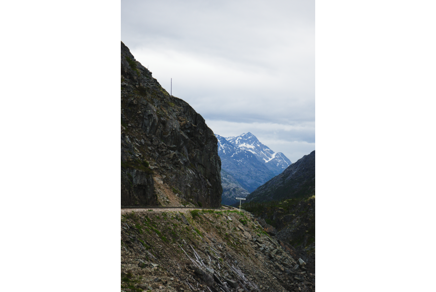 A narrow railway track hugs a rocky cliff edge near Skagway, Alaska, with a sign marking "American Shed." Snow-capped mountains rise in the distance under overcast skies.