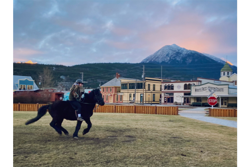 Enjoying cantering through Skagway with historic buildings, snow-capped mountains, and breathtaking sunset in the background.