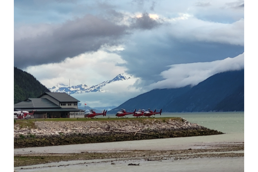 Next to a large two story building with a grey roof and white walls, Five red and white helicopters sit idle on the perfectly level tidelands that steeply giveway to sandy beach in light green colored seas. Ominous clouds hang around the edges of steep dark blue evergreen covered mountains that dive into the fjords in the distance.