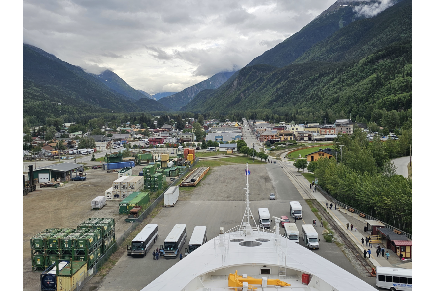 view of downtown skagway from the white bow of a cruise ship features shipping containers to the left and 7 motorcoaches staged to pick up passengers. The white cloud covered skies rest between the evergreen covered steep jagged mountains that give way to the small but flat valley of skagway. Town buildings have a gold rush era style.