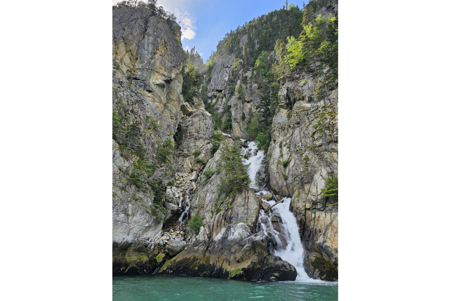 Rocky jagged mountains with scattered green trees on the green water's edge with white frothy waterfall cascading down into the ice cold green sea. Blue sky overhead