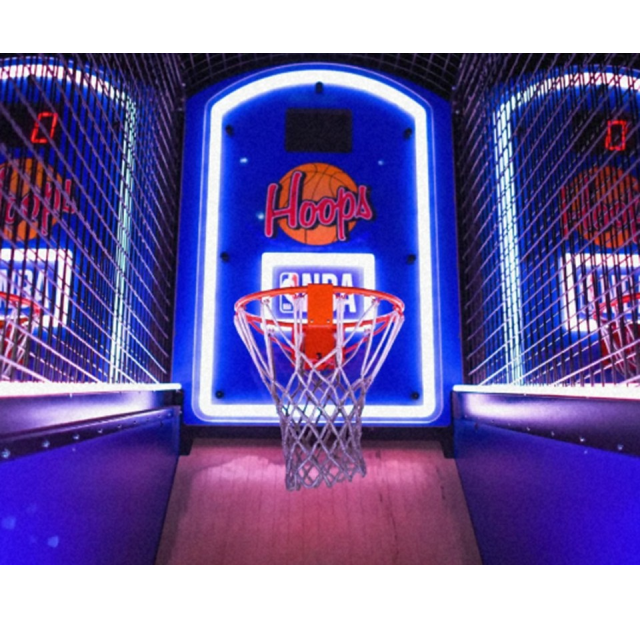 Close-up of a brightly lit basketball arcade game with a glowing blue backboard labeled “Hoops,” an orange rim, and wire side panels inside Sol’s Arcade in Fayetteville, North Carolina.