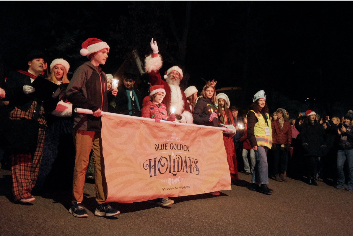 Santa Claus leads the Candlelight Walk 2024 along Washington Avenue, walking in front of a group of participants carrying a festive banner.