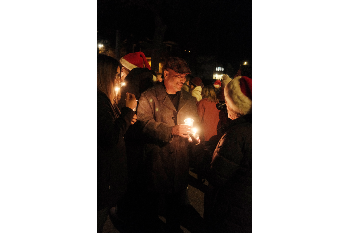 A man with a candle in hand smiles to those around with santa hats on and candles also at night during the Old Golden Holidays Candlelight Walk 2024.