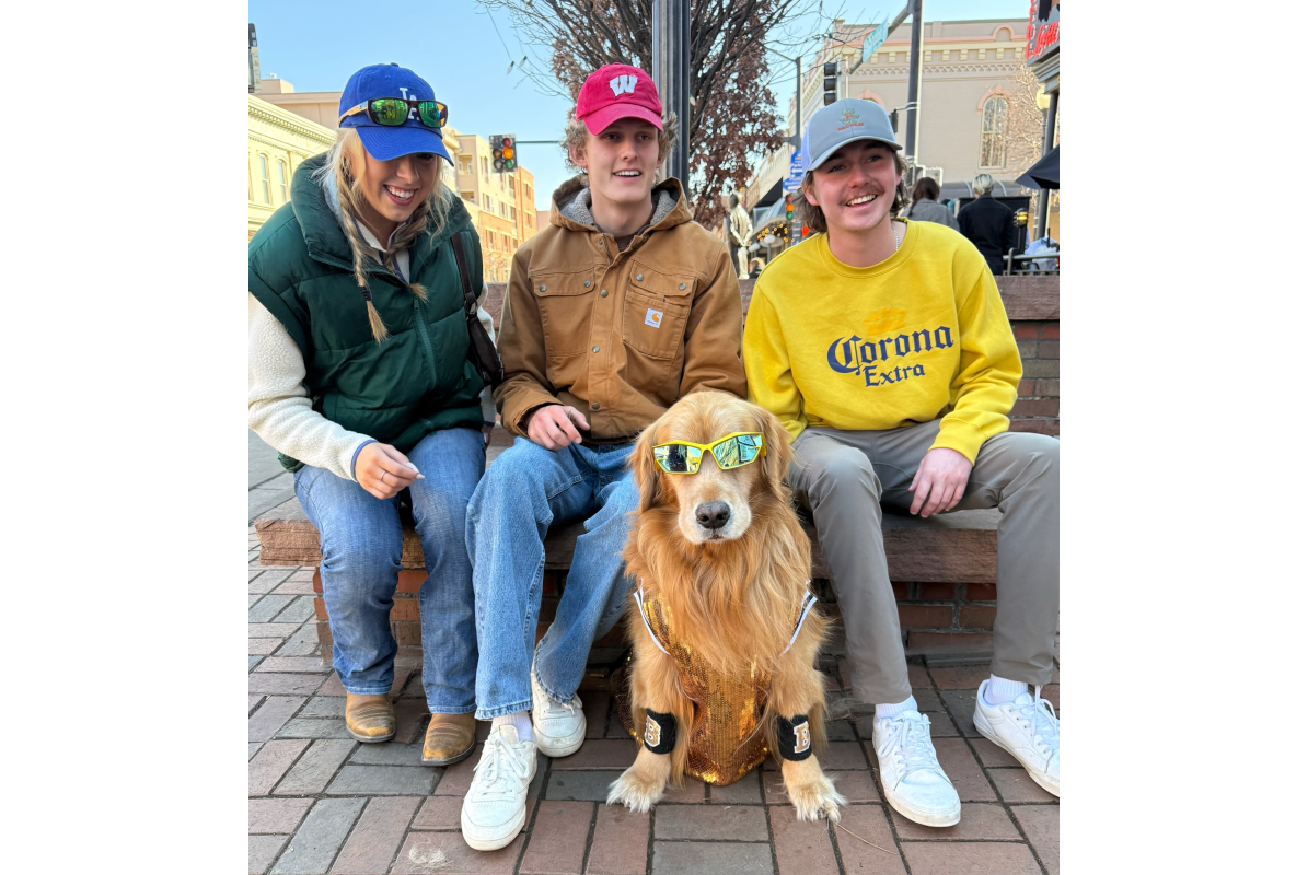 Three people posing with a dressed up and in sunglasses golden retriever at Goldens in Golden CO event 2025