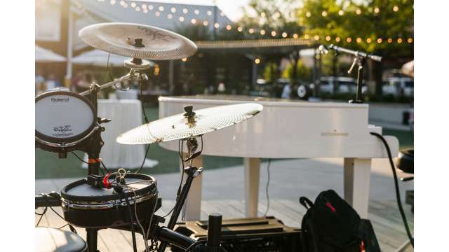 A close-up view of a white piano and black electric drum set. The venue is outdoors, and the sun is setting. There are golden light strands in the background.