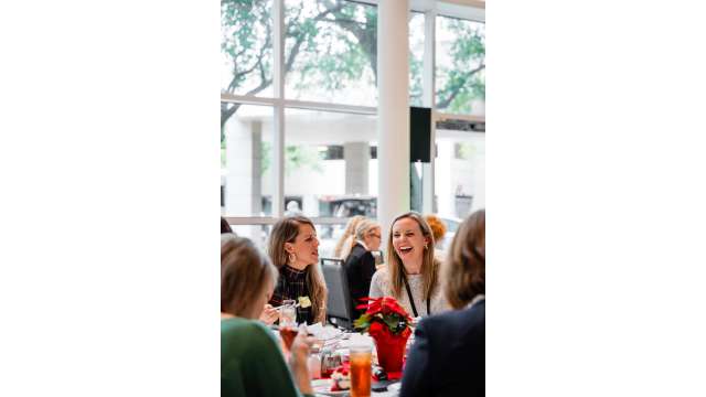 Several women sit at a round table in a room with floor-to-ceiling windows. The two women facing the camera are dressed in business-casual holiday attire (sweaters and red-and-green plaid). The food isn't visible, but there is a tall glass of iced tea and a poinsettia centerpiece.