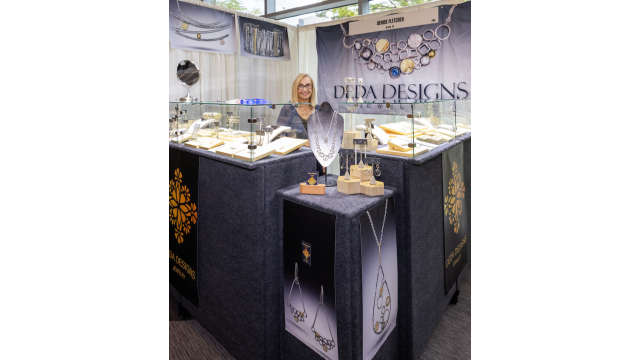 A booth at the Texas Fine Craft Show featuring various display cases of necklaces, bracelets, earrings, and more by Denise Fletcher (a blonde woman with glasses standing behind the counter).